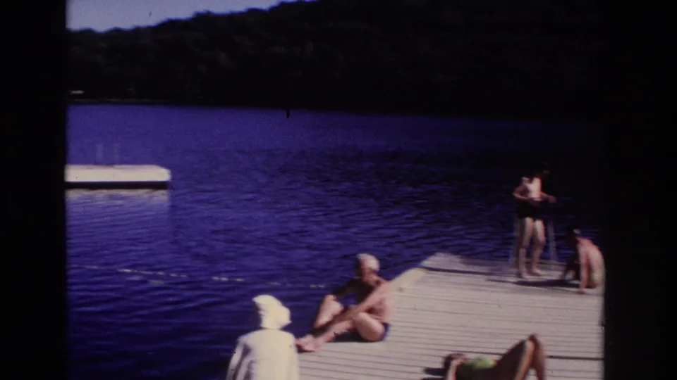 1968 Ocean City home-movie frame with adults in swimwear lounging and standing on a wooden dock by the lake.
