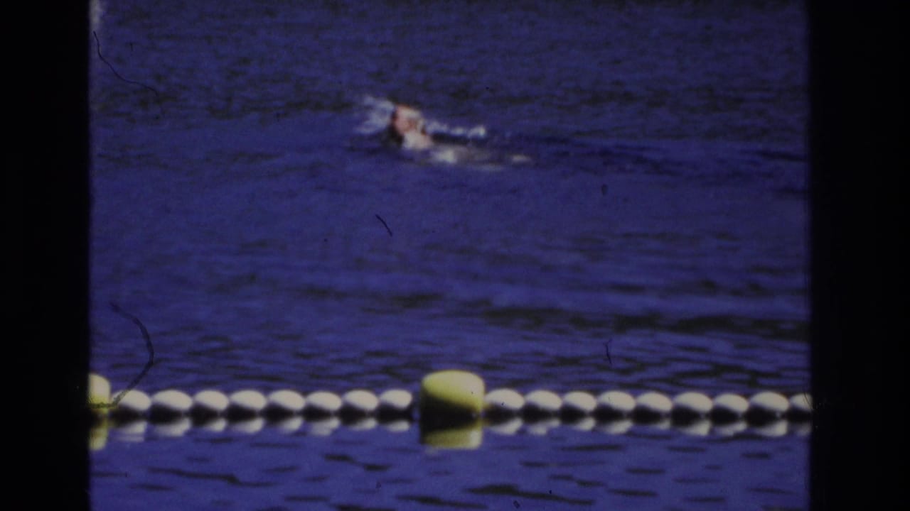 A swimmer is seen near the top of the frame above a buoy line on dark blue water.