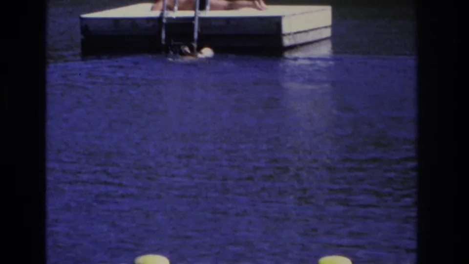 1968 Ocean City home-movie frame of a swimmer beside a floating dock ladder, with a reclining figure partly visible above.
