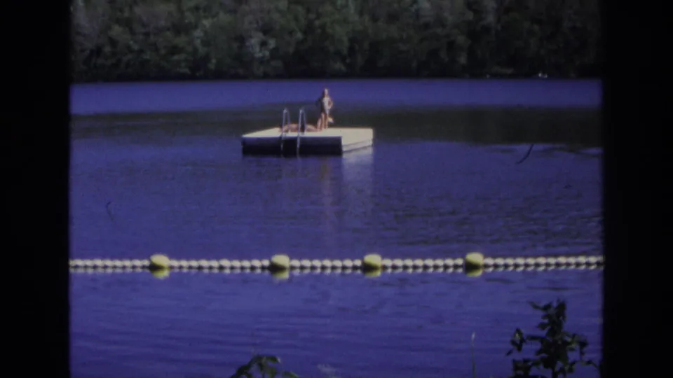 Wide 1968 Ocean City, New Jersey frame with two people on a white floating dock, a buoy line in front, and trees behind the water.