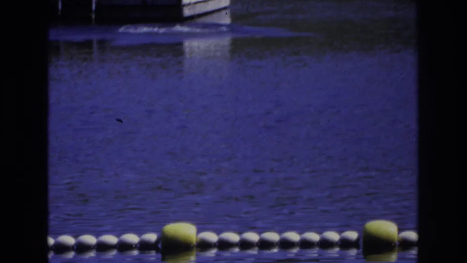 1968 Ocean City home-movie frame showing a buoy-marked swim area seen through dark foreground bars, with rippled blue-purple water.