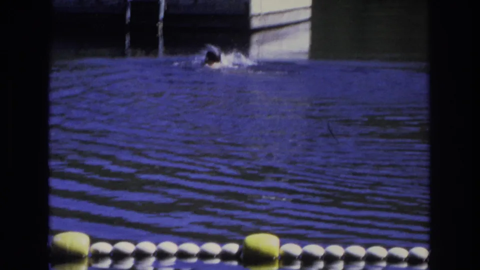 1968 Ocean City frame with a distant swimmer in rippled water, a buoy line in the foreground, and a dock structure behind.