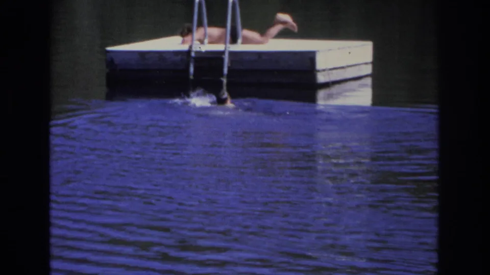 1968 home-movie frame showing a person lying on a floating dock as a swimmer surfaces near the ladder.