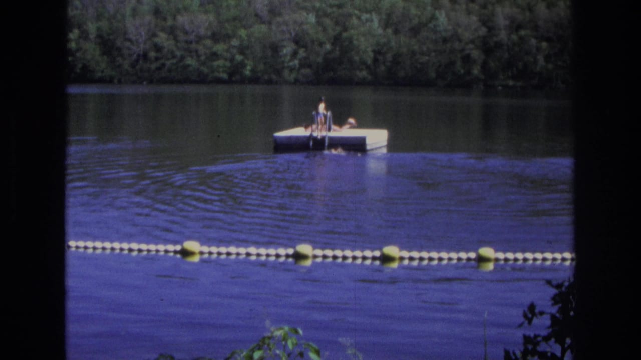 1968 Ocean City, New Jersey home-movie frame showing people on a white floating dock, calm water, buoy line, and wooded shore.
