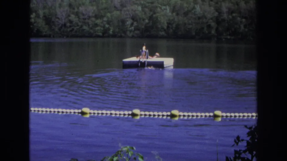 1968 Ocean City, New Jersey home-movie frame showing people on a white floating dock, calm water, buoy line, and wooded shore.