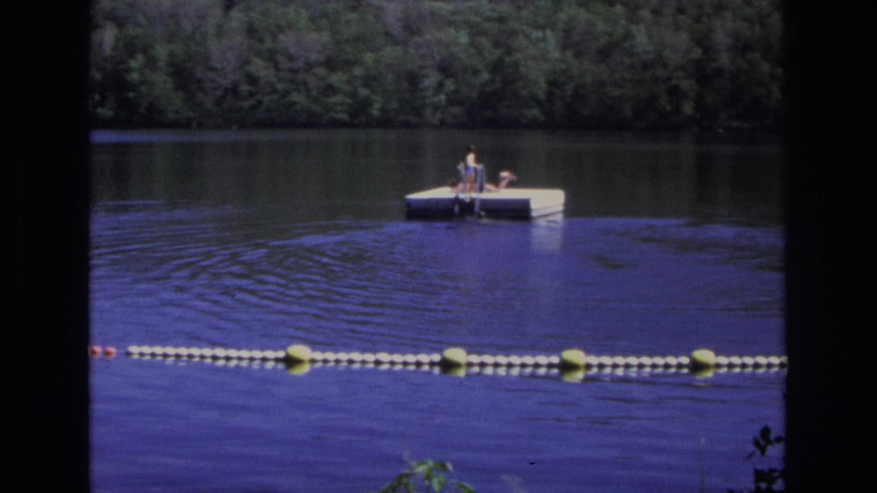 White dock with people near a buoy line, viewed across blue water.