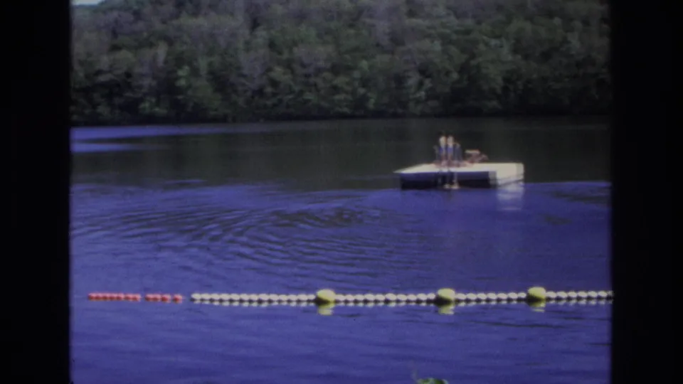 1968 home-movie frame of people on a small white dock across calm water, with a buoy line and wooded shoreline visible.