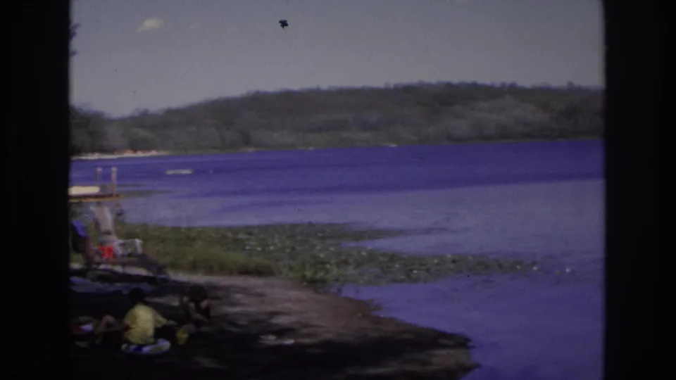 At the end of the pan, a person in a yellow shirt sits near a small dock on the 1968 Ocean City lakeside shoreline.