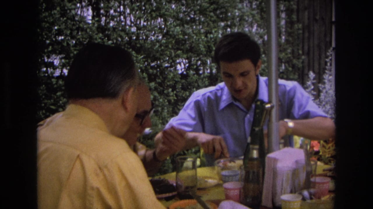 Men dining outdoors, with the man in blue looking down at his plate while others sit in the foreground.