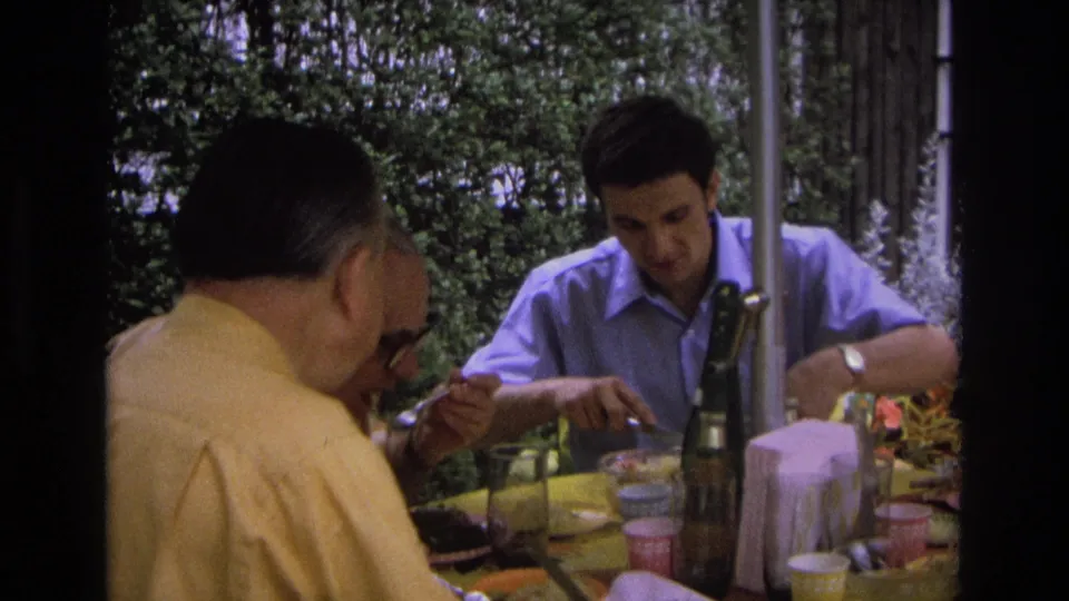 1968 Ocean City home-movie still showing men gathered at an outdoor table, with one partially obscured face and tableware in view.