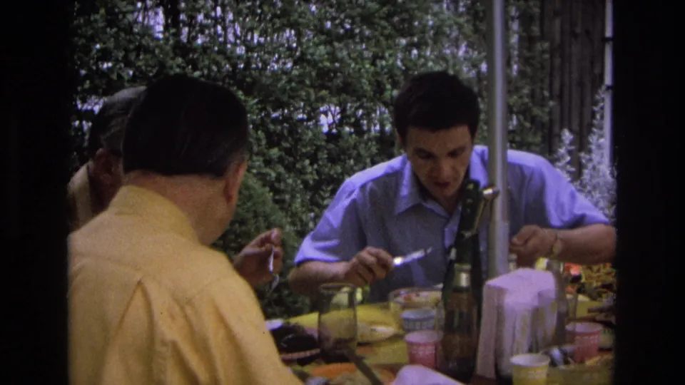 1968 Ocean City frame showing a man in a blue shirt gesturing with a utensil during an outdoor meal with two other adults.