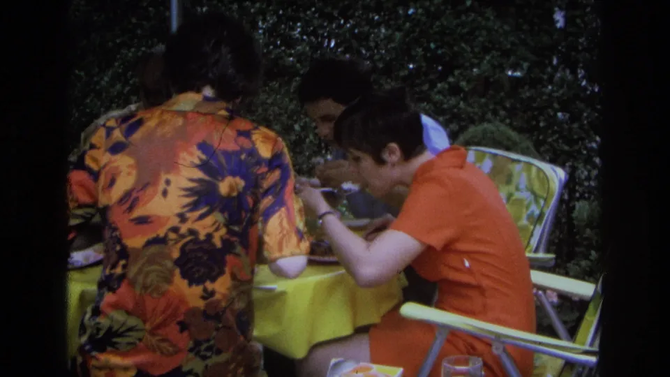 Three adults in 1968 Ocean City sit close together at an outdoor meal, with a yellow tablecloth and foliage in view.