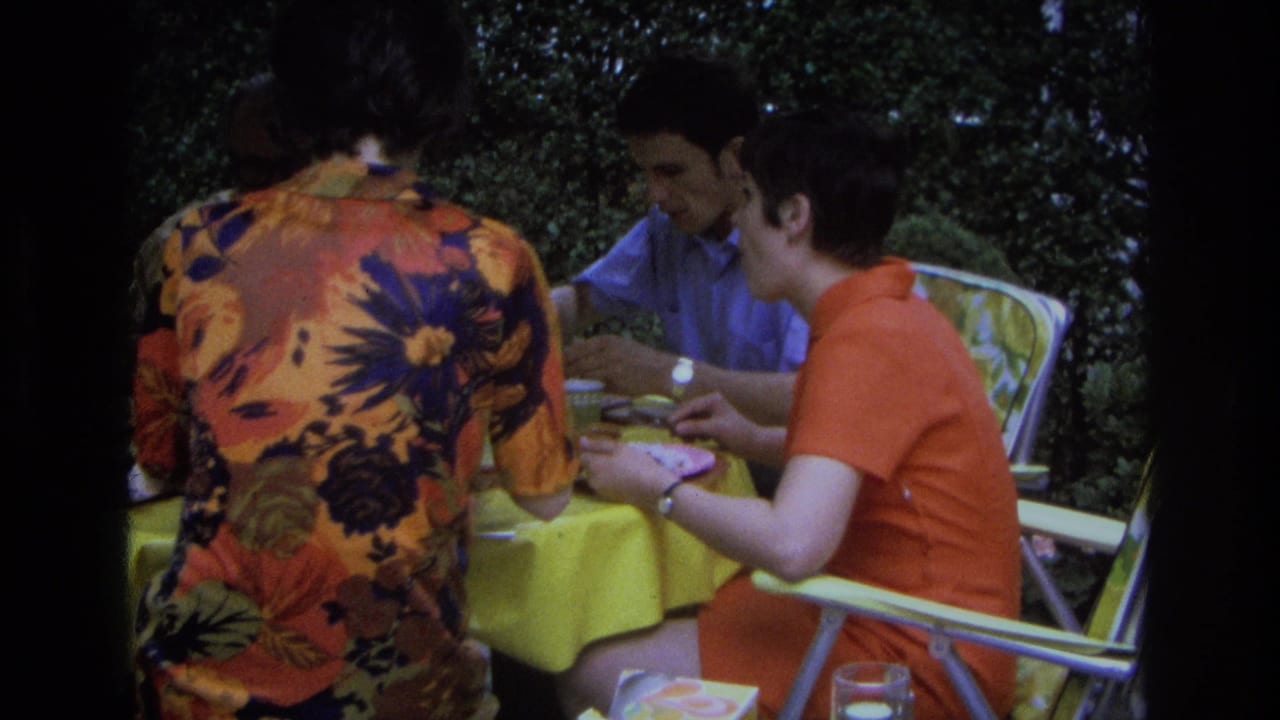 1968 Ocean City home-movie frame showing adults eating and talking at an outdoor table with folding chairs and a yellow tablecloth.
