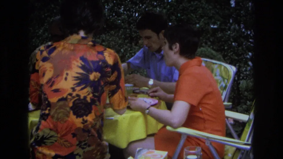 1968 Ocean City home-movie frame showing adults eating and talking at an outdoor table with folding chairs and a yellow tablecloth.