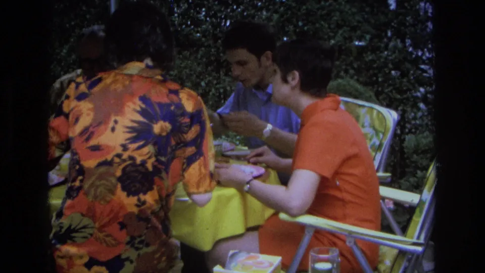 1968 Ocean City home-movie frame with adults seated in folding chairs, eating and conversing at a yellow-covered outdoor table.