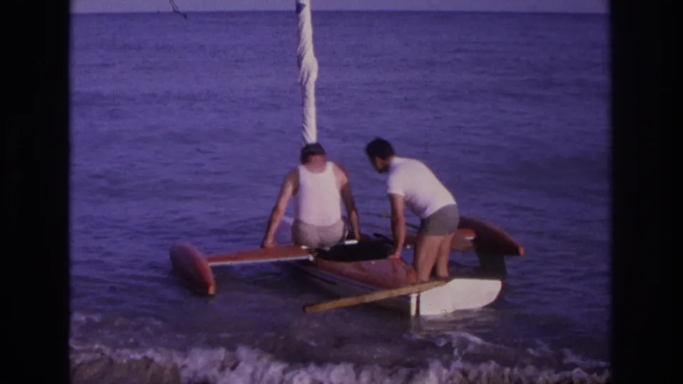 Two adult men work around the red boat in shallow Ocean City surf, with one near the cockpit and another standing at the rear.