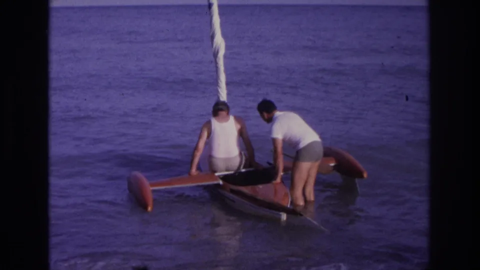 At the end of the Ocean City 1968 clip, the men remain beside the red outrigger-style boat as it floats in shallow ocean water.