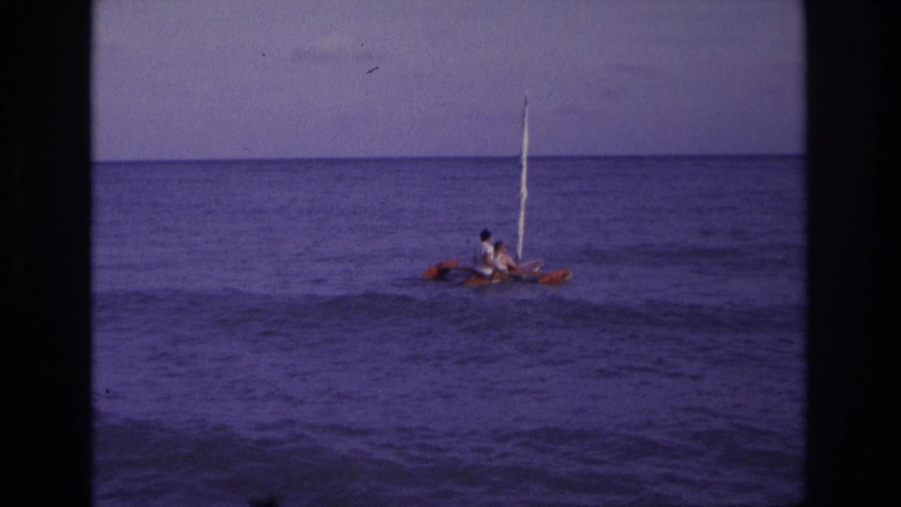 1968 Ocean City, New Jersey home-movie frame showing two distant people on a small red and white sailboat in calm ocean water.