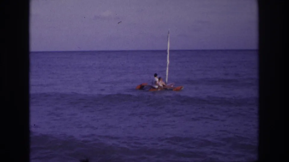 1968 Ocean City, New Jersey home-movie frame showing two distant people on a small red and white sailboat in calm ocean water.