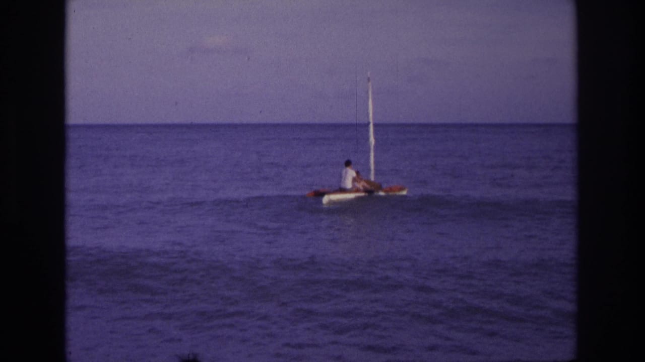 A small mast boat with two people moves across calm ocean water.