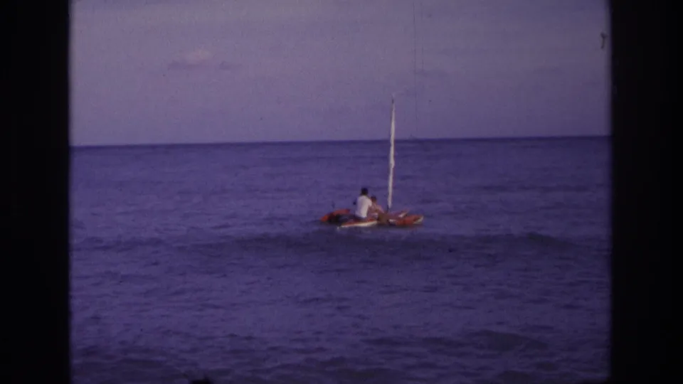 Home-movie frame from Ocean City, New Jersey showing a small sail craft with two distant riders against the ocean horizon.