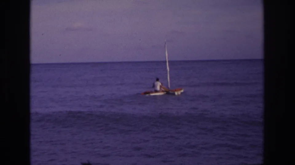 Distant 1968 coastal sailing frame with two people on a red and white small boat moving across purple-hued ocean water.