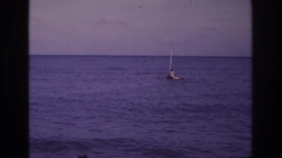 1968 home-movie frame from Ocean City showing a lone person low on a small sail raft in open water.