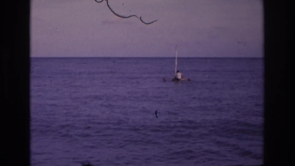 Aged 8mm frame of a distant person on a small sail raft off Ocean City, New Jersey, with film scratches visible near the sky.
