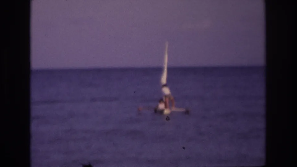 1968 Ocean City home-movie frame showing a man seated in a small sailboat on calm ocean water, with soft focus and faded color.