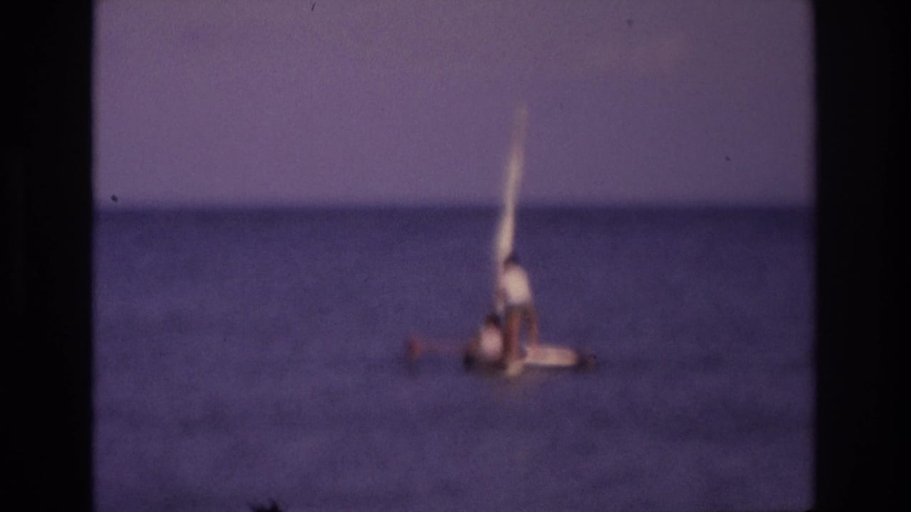 Distant small sailboat tilted on open water with two people aboard.