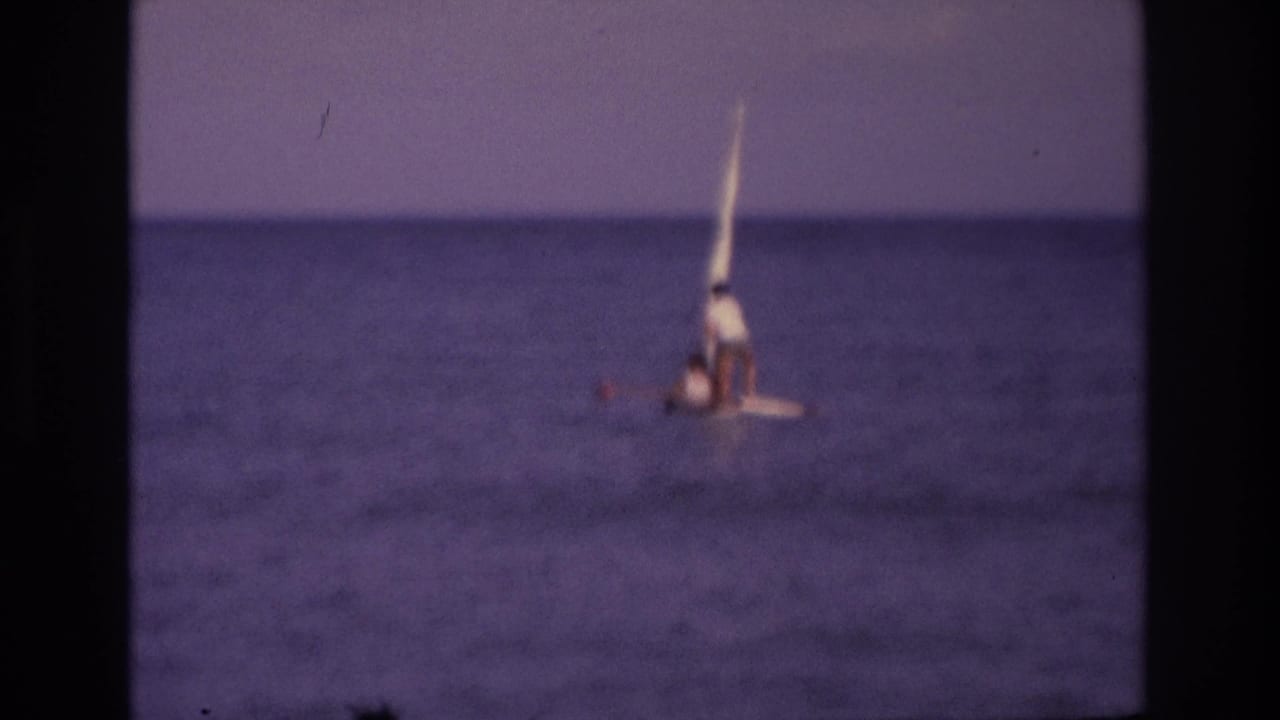 Small sailboat offshore with a high sail and two distant people visible.
