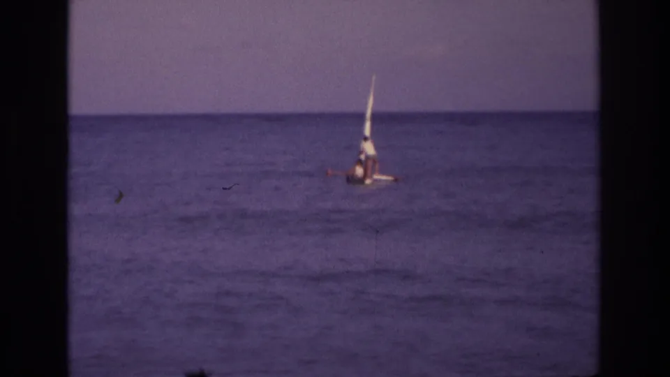 1968 home-movie frame from Ocean City showing the small sailboat leaning offshore, with two distant occupants and visible 8mm film texture.