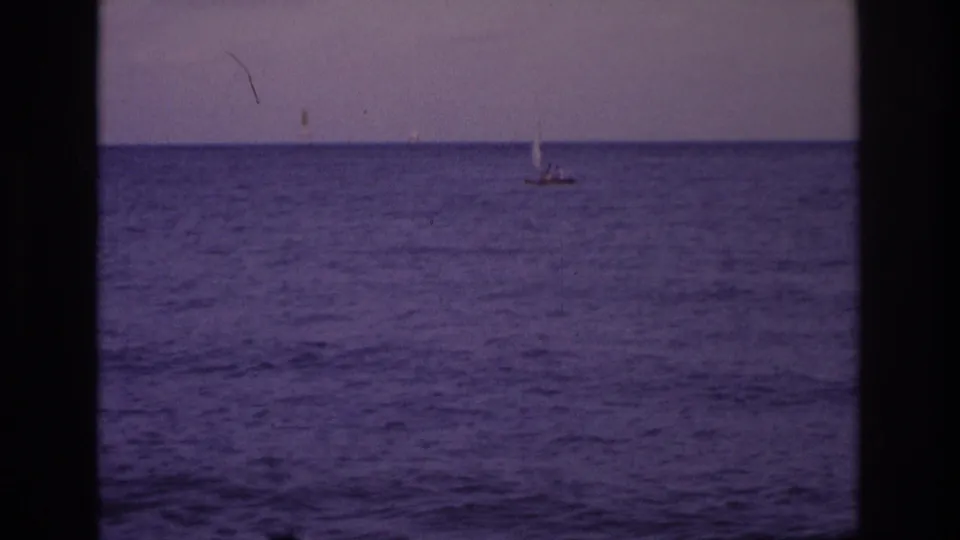 1968 home-movie still of a distant sailboat on purple-blue ocean water, partially framed by dark film or window-like edges.