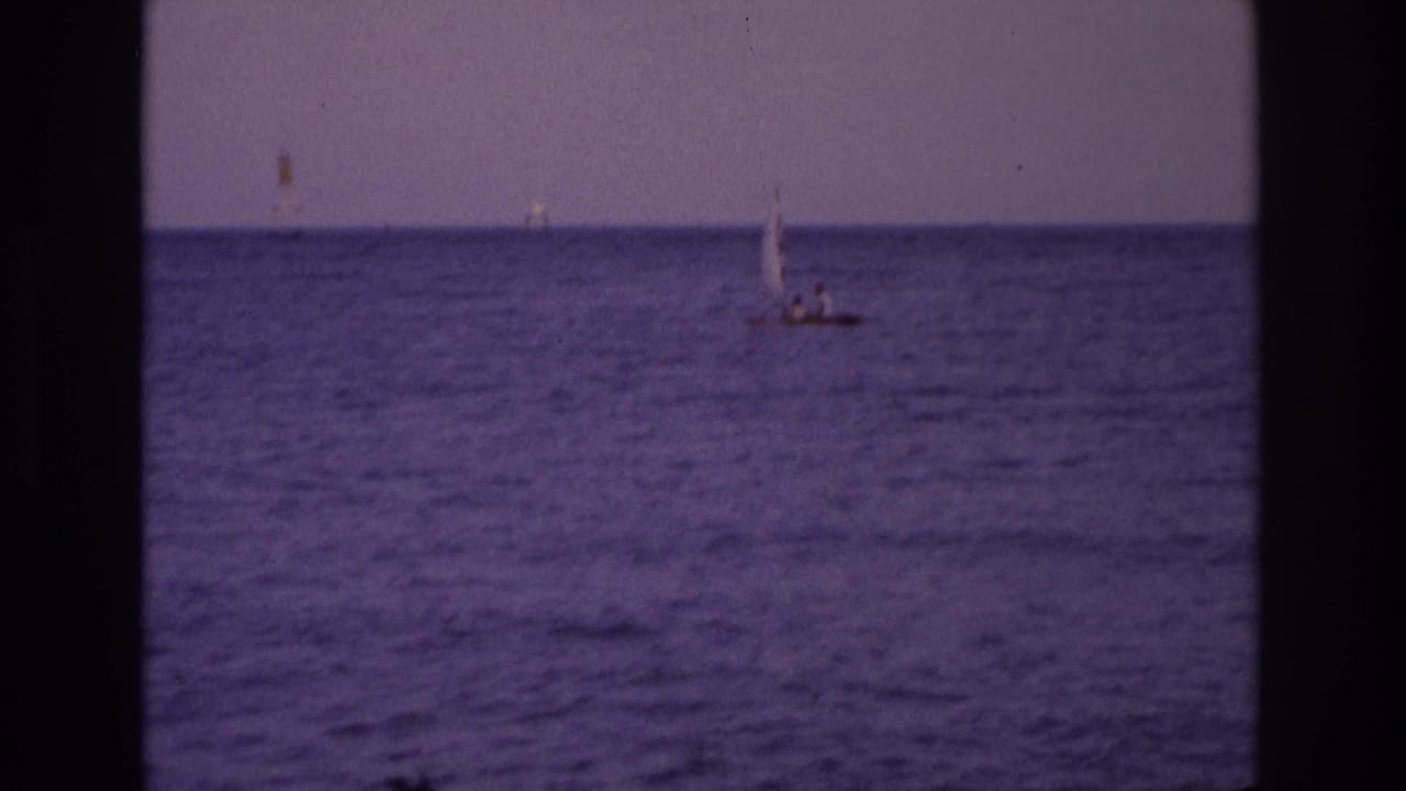 Small sailboat on calm ocean with distant horizon markers and dark film borders.
