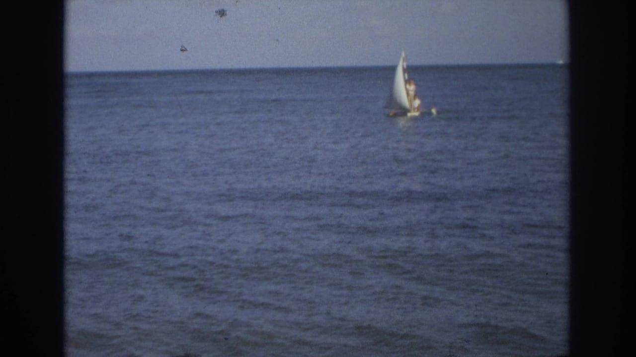 Distant sailboat with a white sail crossing calm ocean water.