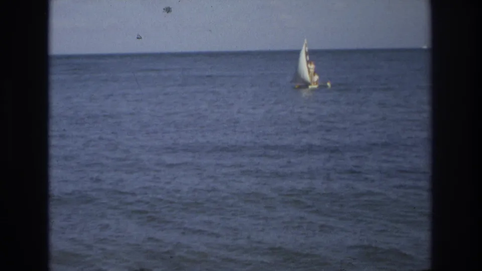 Small sailboat seen across open water in a 1968 Ocean City, New Jersey home-movie frame, with the sailor only faintly visible.