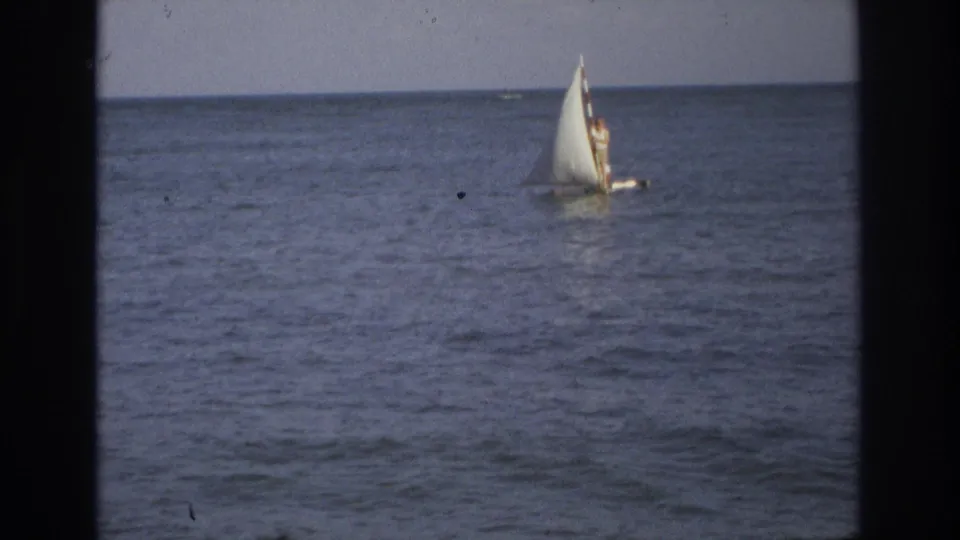 A small sailboat with a faintly visible sailor crosses calm ocean water in 1968 8mm footage from Ocean City, New Jersey.