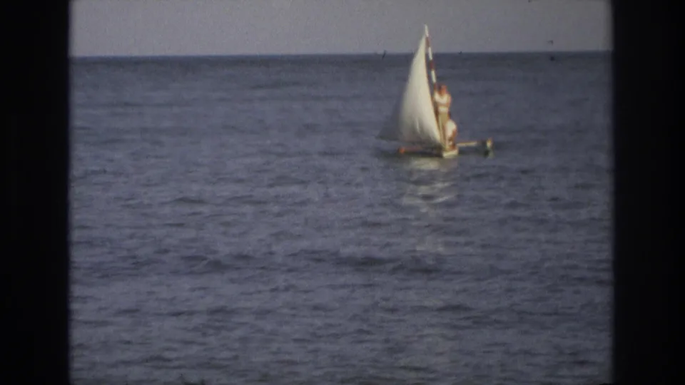 1960s home-movie frame of a small sailboat on the ocean, with one person standing beside the white sail.