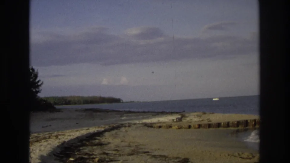 1968 Ocean City, New Jersey beach frame showing sand, low surf, distant shoreline trees, and soft 8mm color fade.