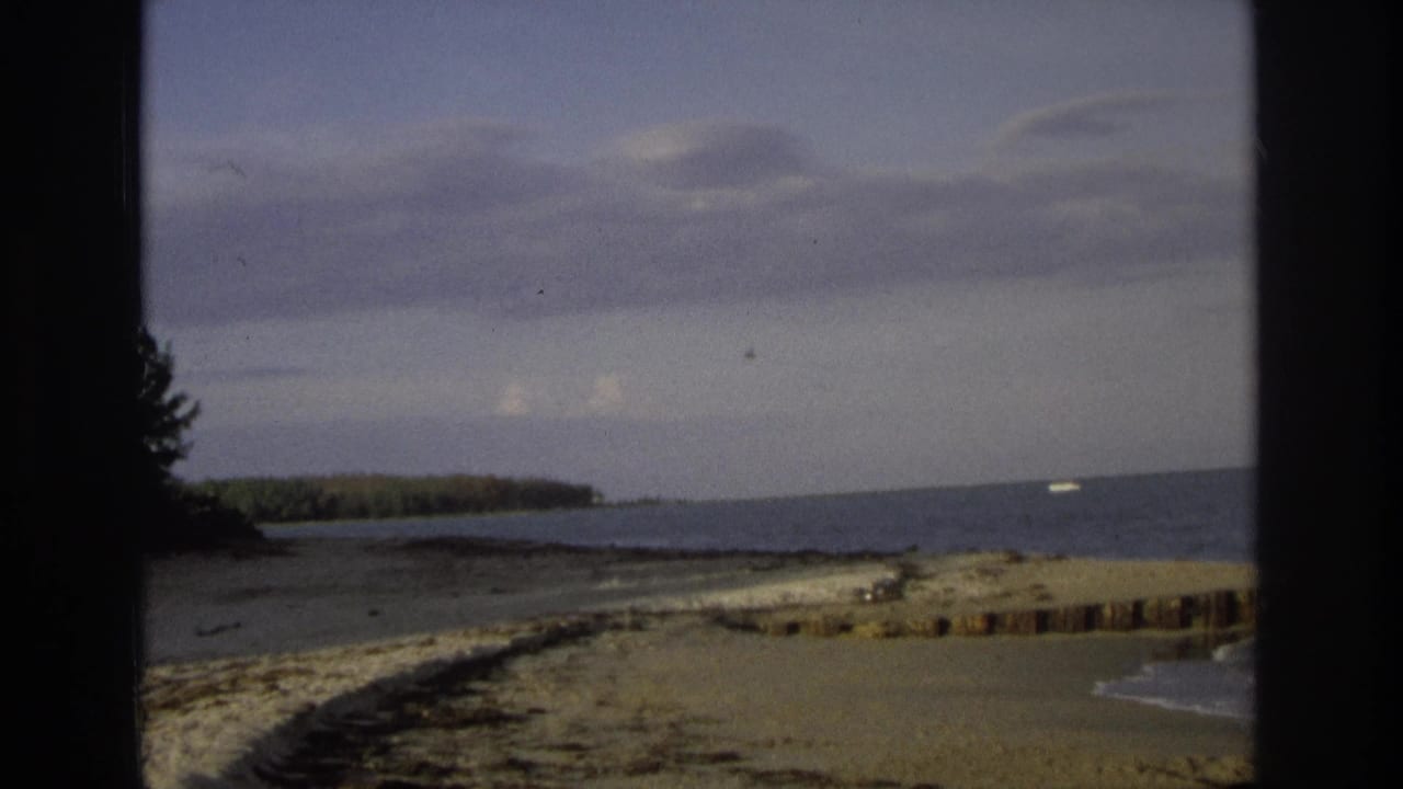 1968 Ocean City, New Jersey home-movie frame showing a cloudy coastal horizon through a dark window frame, with shoreline, trees, ocean water, and a small pale object in the distance.