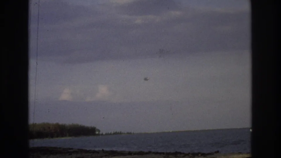 1968 Ocean City, New Jersey frame showing a soft, grainy ocean horizon with a low shoreline, cloudy sky, and a small light-toned object near the right edge.