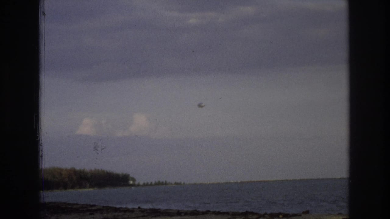 1968 Ocean City home-movie frame showing ocean, low shoreline, cloudy sky, a small aircraft, and a round balloon-like object.