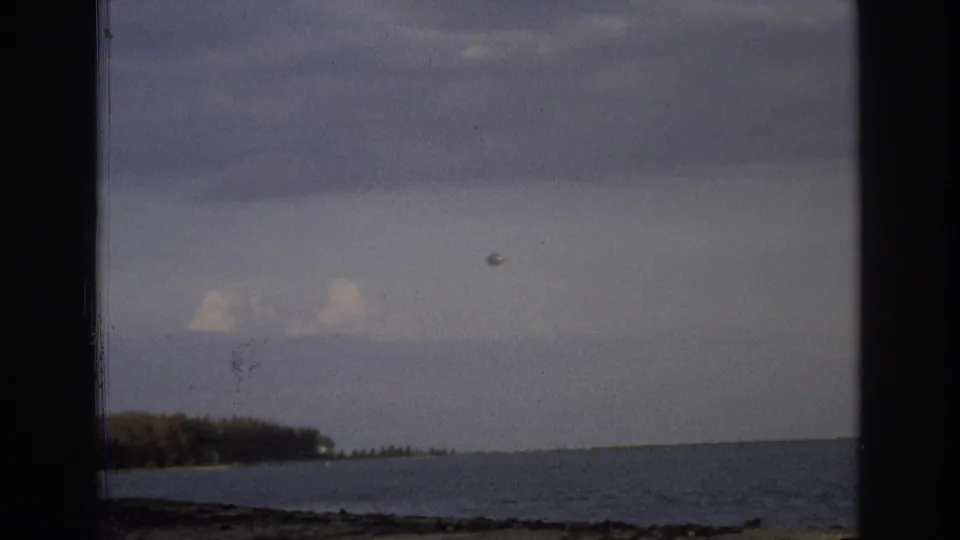 1968 Ocean City home-movie frame showing ocean, low shoreline, cloudy sky, a small aircraft, and a round balloon-like object.