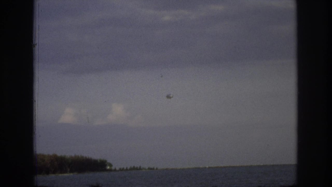 1968 Ocean City coastal frame with a small aircraft and round object in the sky above calm water and a distant treeline.