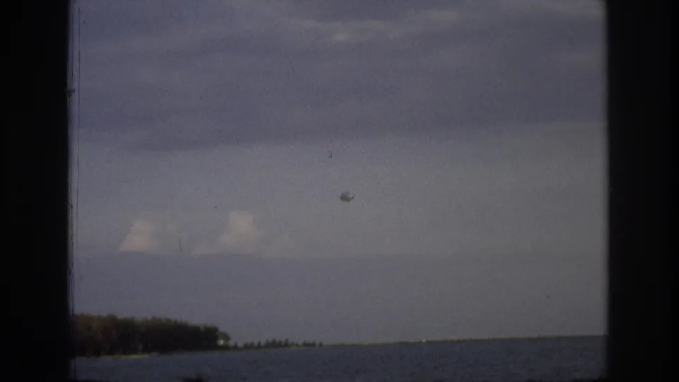 1968 Ocean City coastal frame with a small aircraft and round object in the sky above calm water and a distant treeline.