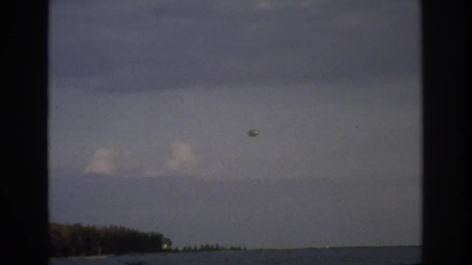 1968 Ocean City 8mm frame showing a low coastal horizon, distant trees, a small aircraft, and a round balloon-like object.