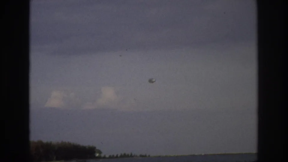 1968 Ocean City, New Jersey 8mm frame showing cloudy sky, calm water, distant trees and a small pale object in the air.