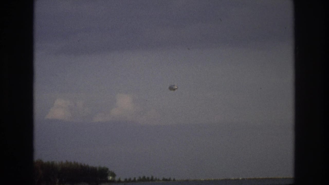 Pale object in cloudy sky above a low shoreline and calm water, framed by dark borders.