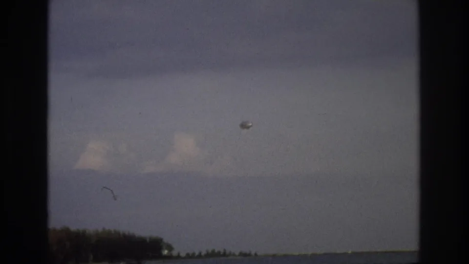 1968 home-movie frame of Ocean City shoreline, showing muted sky, water and a small pale airborne object.