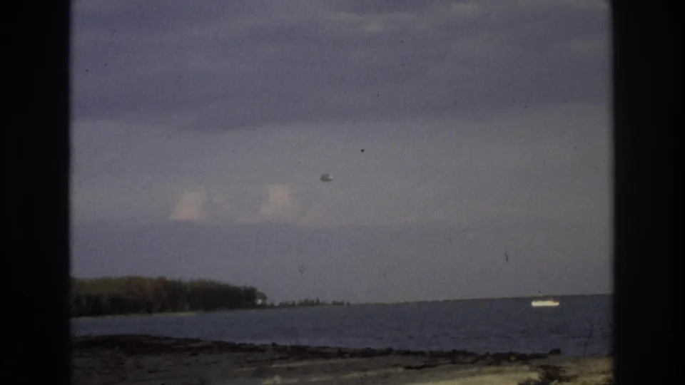 1968 Ocean City, New Jersey shoreline viewed from behind dark frame edges, with a sandy beach, distant tree line, cloudy sky, and small white boat offshore.